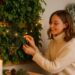 Eine junge Frau in einem cremefarbenen Pullover dekoriert eine Indoor Pflanzenwand mit warmweißen Lichterketten und winterlichen Naturmaterialien, während eine Kerze im Vordergrund gemütliches Licht spendet.