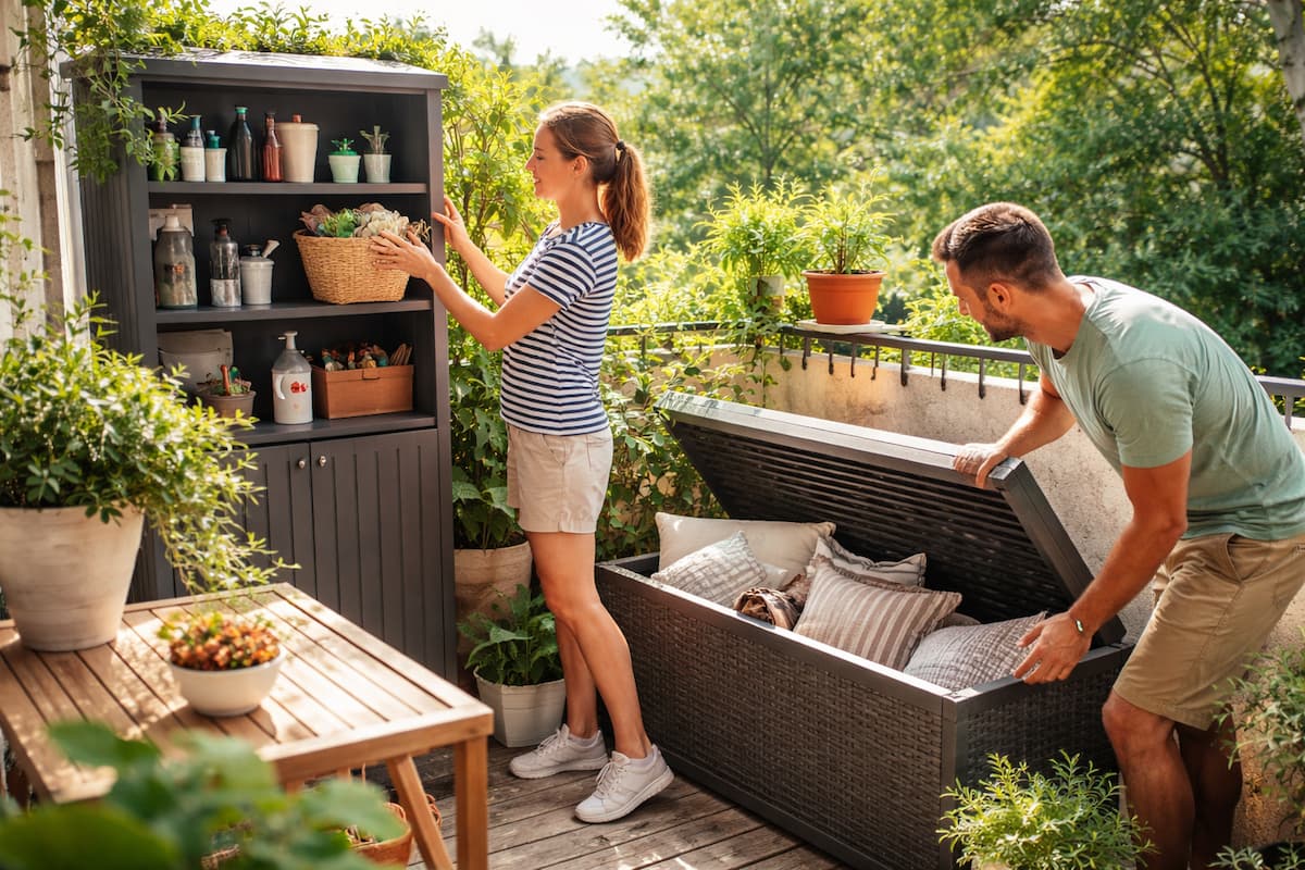 Balkon Stauraum: Paar räumt Kissen in eine große Aufbewahrungsbox und organisiert Gartenzubehör in einem schmalen Balkonschrank auf einem kleinen, grünen Balkon.